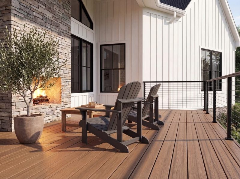 Wood-look deck with a black cable railing, two Adirondack chairs and an outdoor stone fireplace beside a white board-and-batten house